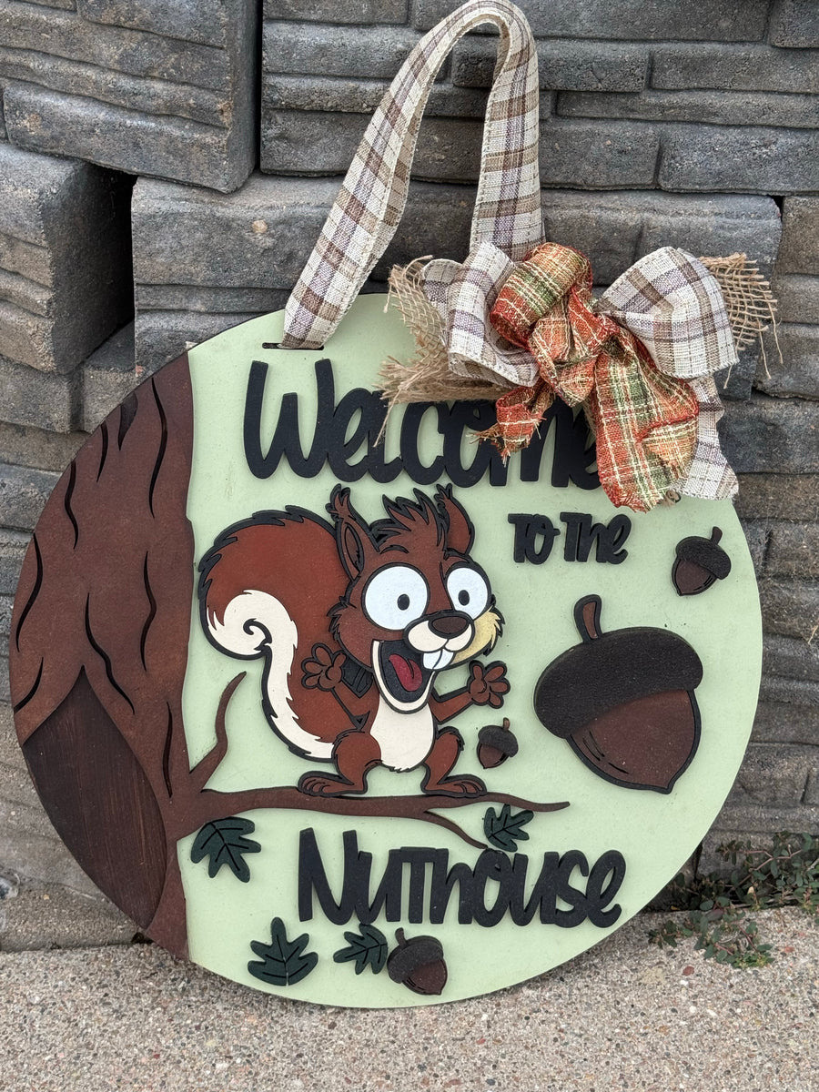 A round decorative wooden sign shaped like a nutshhouse, featuring a cartoon squirrel on a branch, acorns, and the words 'Welcome to the Nuthouse.' The sign has a pale mint green background, dark brown lettering and tree trunk details, plaid fabric hanging straps, and a large plaid bow at the top.