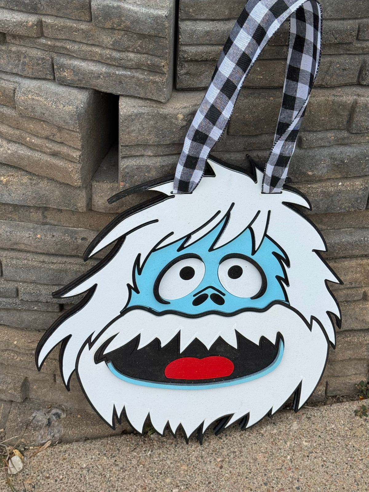 A cartoonish yeti face hanging decoration with a black and white gingham ribbon handle, set against a gray stone wall background.