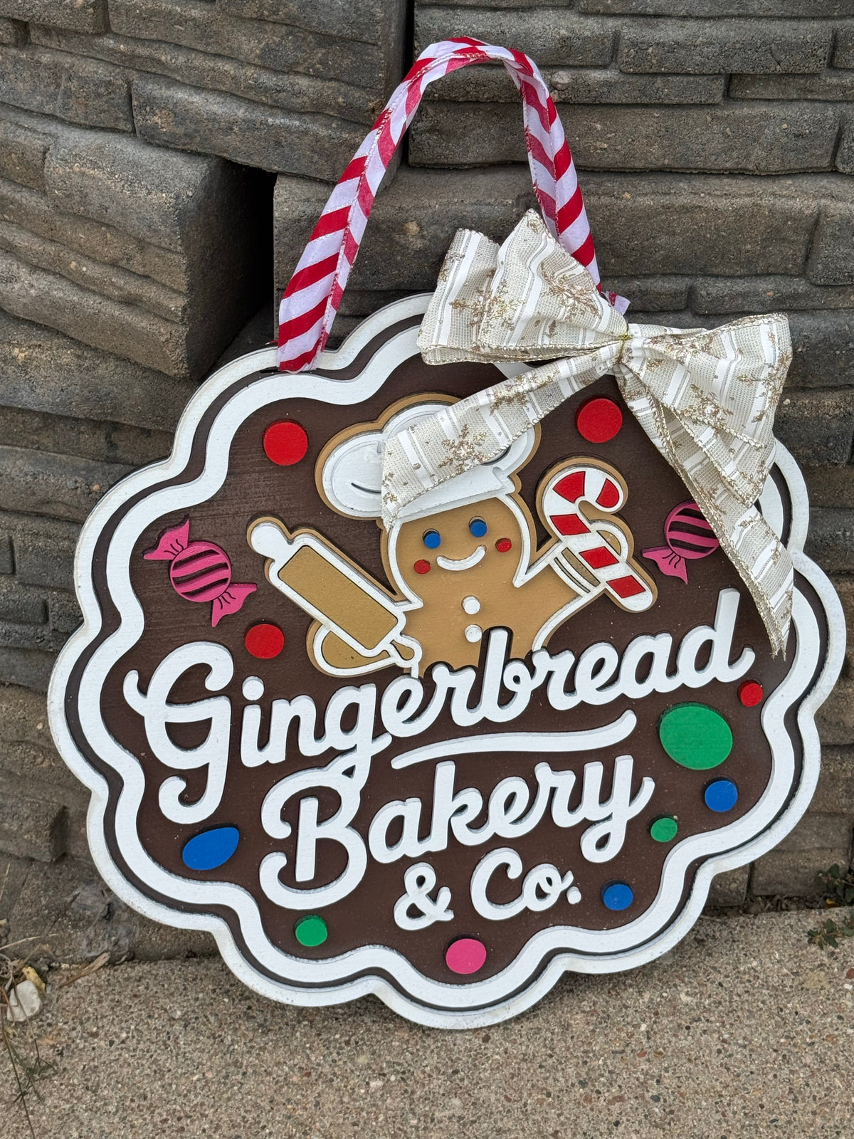 A scalloped gingerbread-themed hanging sign that reads 'Gingerbread Bakery & Co.' in white script on a brown background, decorated with a gingerbread cookie, candy cane, peppermint, and colorful dots.