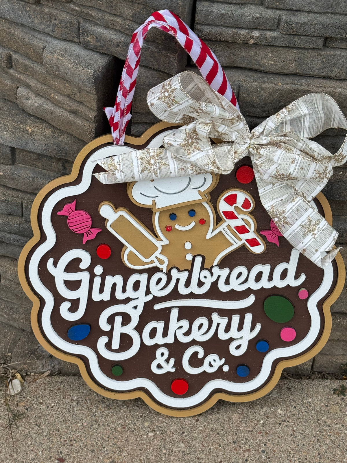 A gingerbread-shaped decorative sign reading "Gingerbread Bakery & Co." with a white bow and striped hanging ribbon, photographed against a stone wall.