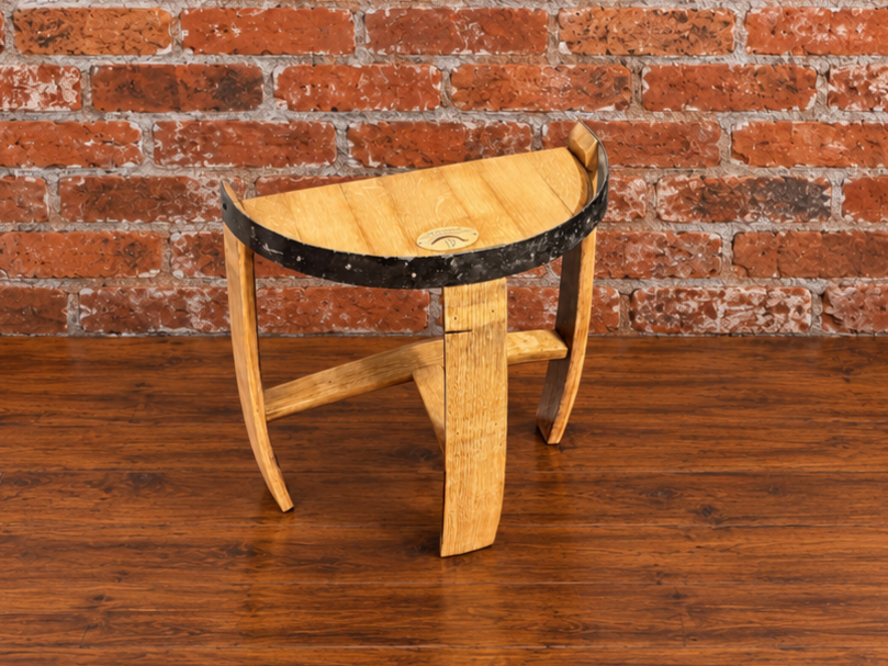 A rustic wooden stool with a semi-circular seat, made from reclaimed wood pieces including a barrel band, set against a red brick wall and dark wood floor.